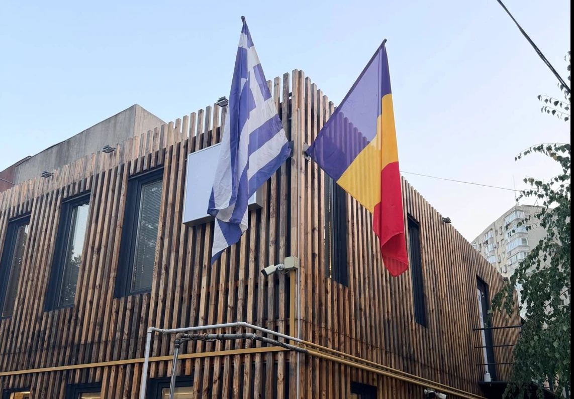 Fațada magazinului Bunătăți din Grecia de pe Strada Chiparosului 34: clădire îmbrăcată în șipci de lemn, cu steagul Greciei și steagul României la etaj